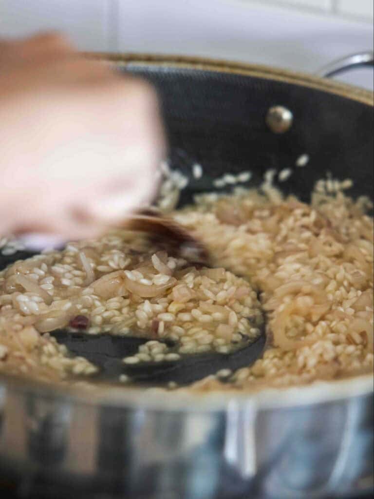 A close-up of a person stirring creamy risotto in a black pan on the stove, with visible grains of rice and caramelized onions.